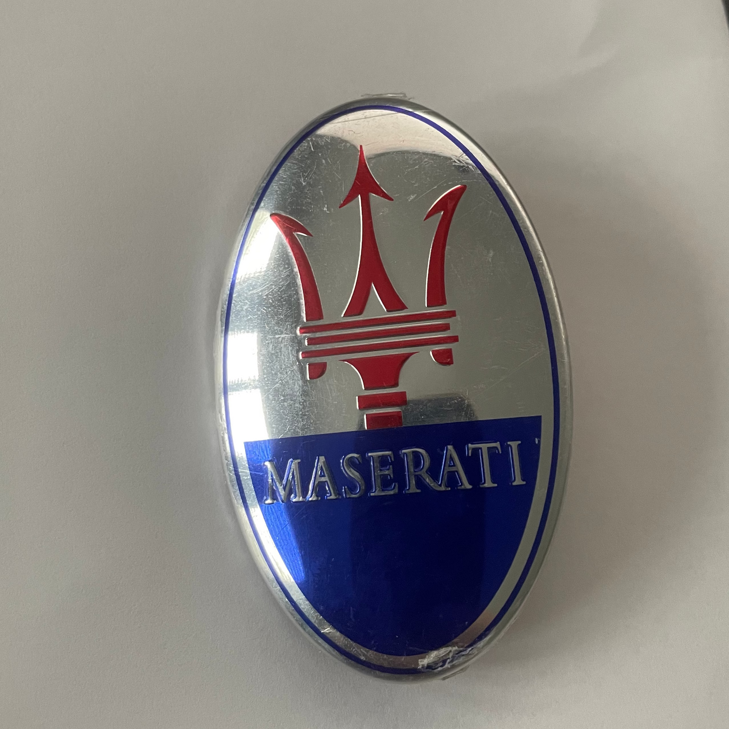 An oval Maserati emblem badge featuring the red trident logo on a silver background with a blue band reading MASERATI, photographed on a white surface.