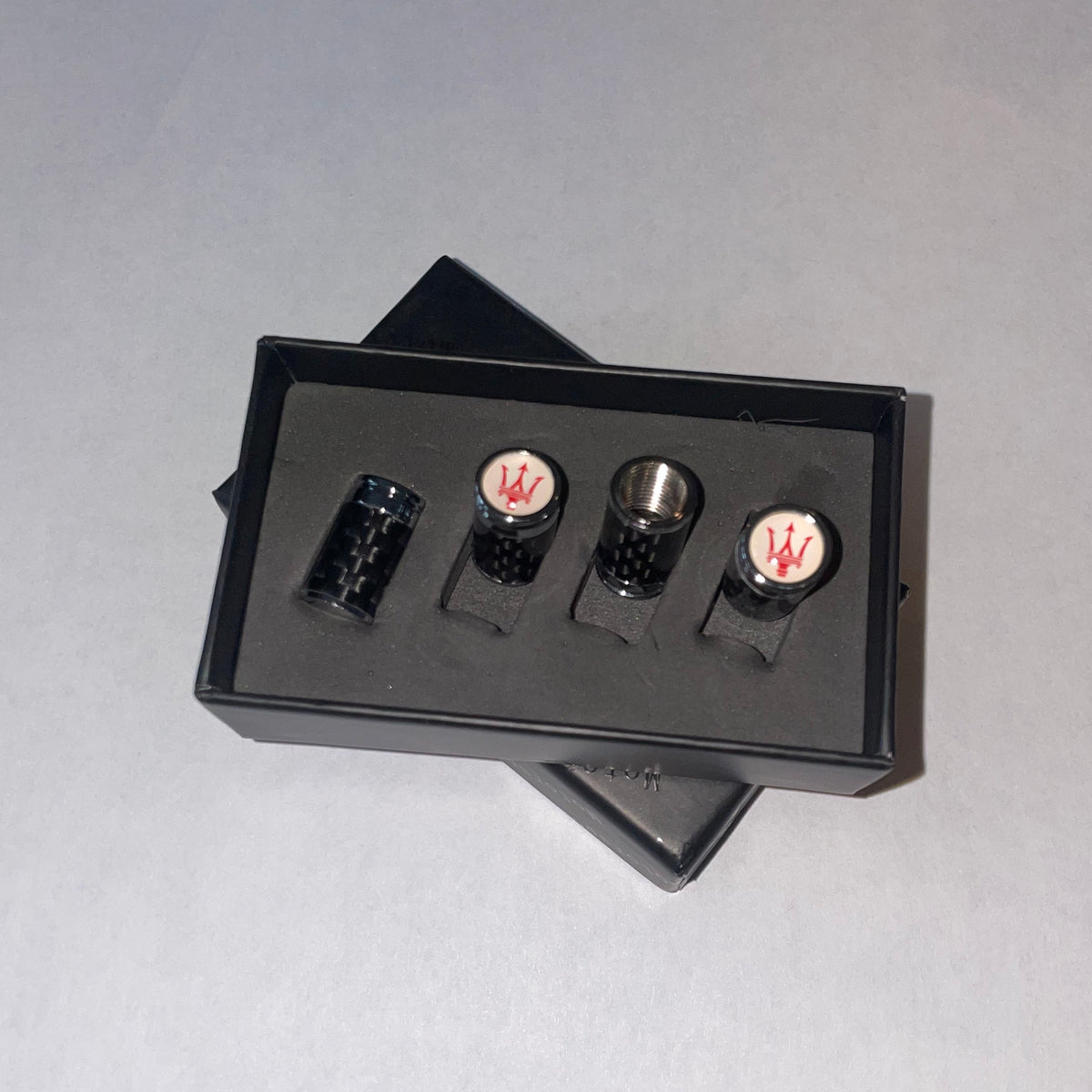 A set of four black cylindrical accessories, likely lip glosses or small bottles, displayed in a black presentation box. Two of the items feature metallic silver caps with a red stylized 'W' logo, and two have black caps.
