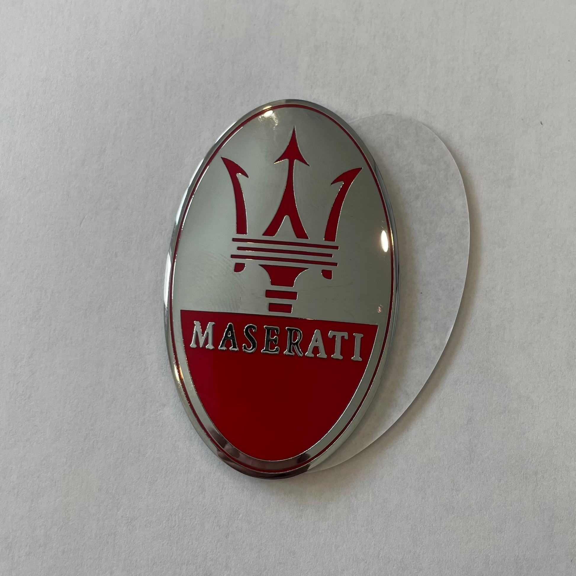 An oval Maserati emblem badge featuring the red trident logo and the brand name 'MASERATI' on a red and silver background, shown on a light gray textured surface.