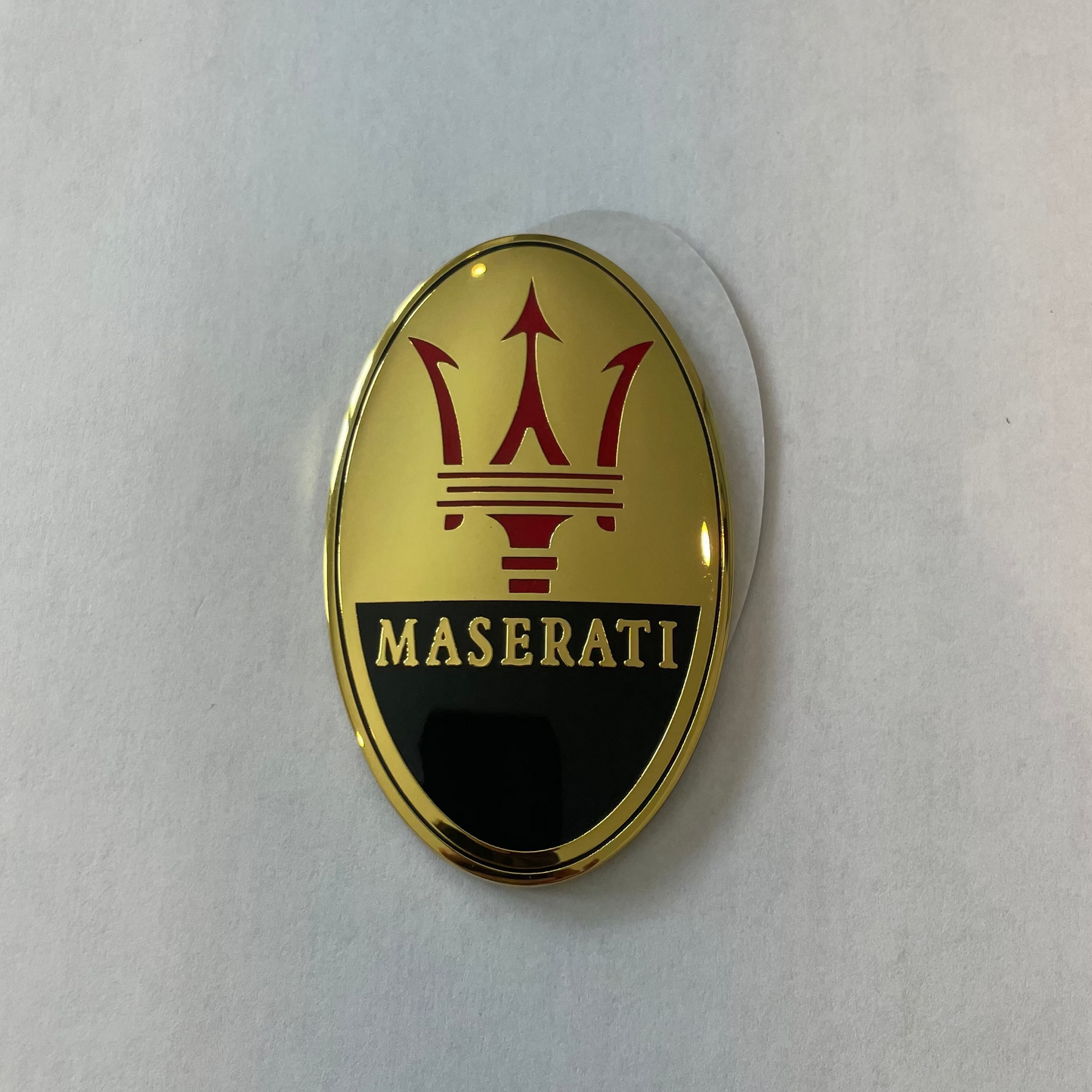 An oval Maserati emblem badge featuring the brand's trident logo in red on a gold background with a black lower section and gold lettering, shown on a light gray surface.