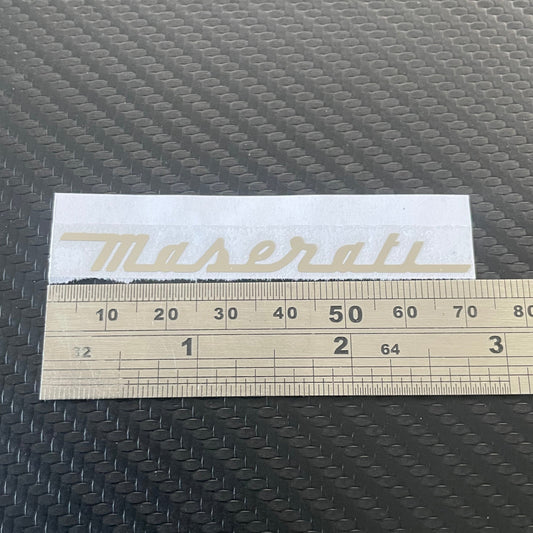 A rectangular white decal with the Maserati script logo in gold, shown next to a wooden ruler on a dark textured surface.